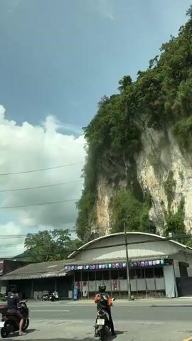 The video shows a roadside scene with a large, sheer cliff face covered in lush green vegetation on the right. In the foreground, a paved road runs horizontally across the frame. On the left side of the road, there are several small buildings with peaked roofs, some with awnings and what appear to be shopfronts. A tall utility pole with multiple wires stretches upwards near the buildings. To the right of the road, a motorcycle is parked. The sky above is partly cloudy with a bright, daytime light. The second clip pans slightly to the right, revealing more of the cliff face and a street light.