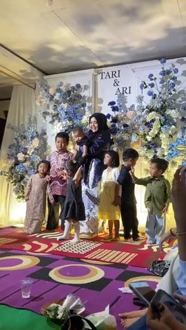 This media file captures a heartwarming scene at an event, likely a wedding reception given the backdrop featuring the names "TARI & ARI" in large, elegant lettering. The setting appears to be indoors, illuminated by warm artificial lighting. A woman, dressed in a dark hijab and a beautiful patterned outfit, stands in the center, holding a baby. She is surrounded by six children of various ages, all dressed in festive attire.

The children are positioned in front of the woman, some standing close and others slightly behind her. The youngest child is being held by the woman, while the others are on their own. They appear to be posing for a photograph. The atmosphere seems joyful and celebratory, with the woman smiling warmly at the camera. The children also display a range of expressions, with some looking at the camera and others engaged in their own world.

The backdrop is adorned with abundant floral arrangements in shades of blue and white, interspersed with lighter-colored flowers, creating a visually appealing and romantic ambiance. A white curtain is visible on the left, and a plush, multi-colored carpet with a circular pattern covers the floor in the foreground. On the carpet, there are some discarded plastic cups and wrappers, suggesting that the event is ongoing or has recently concluded. The overall impression is one of family, togetherness, and a special occasion being celebrated. The time of day cannot be determined from the image, but the indoor setting and lighting suggest it could be evening. The location context of Kembang Harum, Indonesia, adds a cultural element to the scene.