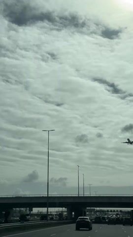 From a vehicle driving on a highway under an overpass, a wide-bodied jet is seen in flight under a cloudy sky with a glimpse of the sun. Several vehicles are visible on the highway.