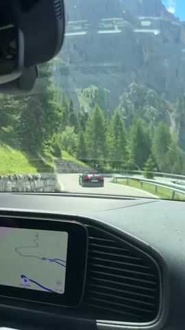 The video shows a view from inside a car driving on a winding mountain road. The driver's dashboard and navigation screen are visible in the foreground. The road is paved and bordered by a stone wall on one side and trees and steep mountain slopes on the other. Several sports cars are seen driving on the road ahead and in the opposite direction. The sky is blue with some clouds, and the overall environment appears to be a sunny day in a mountainous region with abundant green vegetation.