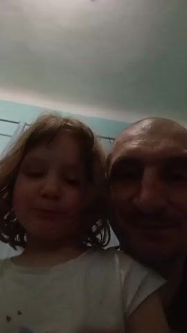 The video shows a close-up of a man and a young girl. The man has a shaved head and is smiling slightly. The girl has light brown hair and is wearing a white t-shirt with a design on it. They appear to be taking a selfie together. The background includes a light blue wall and a curtain rod with sheer curtains.