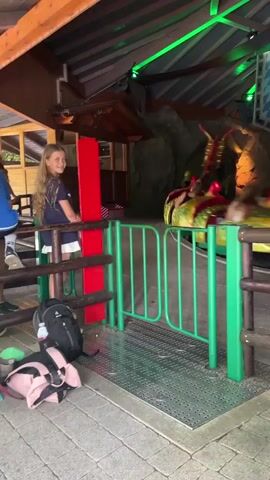 The video shows an amusement park ride with a dragon-themed rollercoaster. A young girl with blonde hair is standing on the left, wearing a dark blue shirt and white skirt. She is looking towards the camera and smiling. In front of her, a black backpack and a pink bag are on the ground. A green fence separates her from the ride. The rollercoaster, designed to look like a dragon, is moving on a track and is occupied by several people. The ride is indoors, with a wooden roof and rock-like decorations on the walls. Green lights illuminate the area. A flag with a lion emblem is hanging on the wall. Another child, wearing a light-colored shirt with a pattern, is standing on the right side of the frame, watching the ride. The ground is paved with gray stones, and a metal grate is visible near the fence. The rollercoaster is brightly colored with red and yellow accents.
