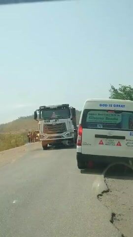 The video shows a view from inside a car of a truck pulling away from a white passenger van on a paved road. The white truck is labeled “OGB” and “GABRIEL” on its front. It is hauling a load covered by a dark tarp. The road is cracked, and there is dry vegetation along the sides. The white van has a sign reading “GOD IS GREAT” and “Explore West Coast With Us Ghana Route” and includes contact information. The van is labeled “B25.” The view suggests a sunny day with a hazy sky.