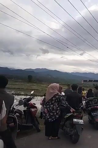 The video shows a cloudy day with a rural landscape featuring rice paddies and mountains in the background. Several people are gathered around a white car that appears to be stuck in the mud. Motorcycles are parked nearby. In the later part of the video, a yellow crane is visible, suggesting an attempt to extract the car. Some individuals are observing the situation, while others are interacting with the car or the crane. The overall atmosphere seems to be one of concern or problem-solving.