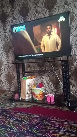 The video shows a television in a room. The screen is displaying a movie trailer for "Dushman aka-uka: kinopremyera." The TV is placed on a black glass-top stand. On the stand beneath the TV, there are several items including a brown paper bag, a colorful bucket with Disney characters, and a pair of pink plastic baby shoes. In front of the TV stand, there is a patterned red, brown, and white blanket. The wall behind the TV is covered in patterned wallpaper. Throughout the video, the scenes on the TV screen change, showing both close-ups of actors and wider shots of a crowded event.