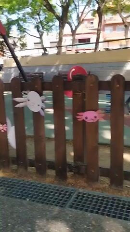 The camera pans right along a wooden playground fence. The fence is made of rounded vertical posts, and decorated with wooden fish and crab shapes, painted in colors like pink, purple, blue, and brown. The playground inside the fence has a mulch surface. Outside the fence is a metal storm drain. In the background are houses and trees. People can be seen walking in the background. The day appears to be sunny.