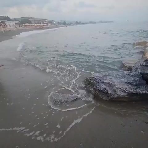The video shows a close-up of a wave crashing on a rocky beach. The water is gray and choppy, with white foam washing over the dark rocks. In the background, a long stretch of beach is visible, lined with buildings and umbrellas. The sky is overcast.