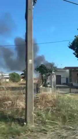Here is a detailed description of the video:

The video shows a large plume of dark smoke rising into a clear blue sky. The smoke appears to be coming from an area behind some buildings and vegetation. The camera is moving, likely from a vehicle driving along a road. The foreground consists of dry grass and some plants. A two-story building with white walls and red trim is visible on the left side of the frame. There are power lines stretching across the top of the video. The overall impression is of a fire or some other event causing a significant amount of smoke.