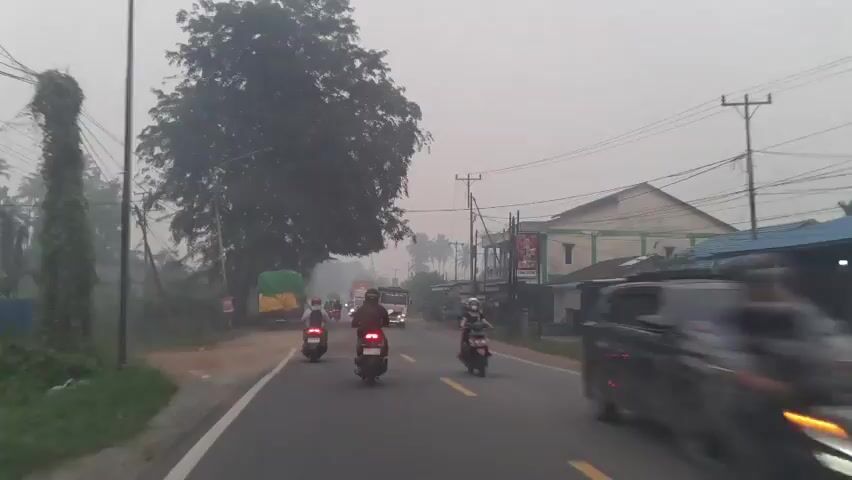 A vehicle is driving down a road, likely in Southeast Asia. The road is paved and has yellow dividing lines. There are several other vehicles on the road, including motorcycles, cars, and trucks. The trucks are carrying large loads, some covered with tarps. There are buildings on the side of the road, some appearing to be shops or homes. There are also trees and utility poles visible. The sky is overcast, and there appears to be some haze or smoke in the air, suggesting possible air pollution or fog. The overall impression is of a busy, developing area with ongoing traffic.