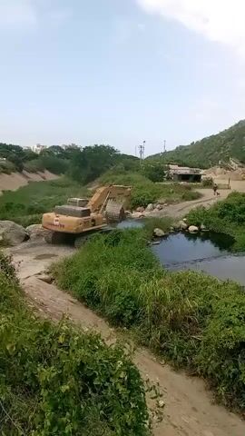 A yellow excavator is shown operating in a shallow body of water, possibly a creek or a stream. The excavator has its bucket submerged in the water and appears to be digging or clearing material. The surrounding environment is lush with green vegetation, including tall grasses and bushes. There are some rocks visible in and around the water. In the background, a hillside covered in greenery and some buildings can be seen. The sky is overcast. The excavator has "KOMATSU" written on its side, along with a model number that appears to be "400".