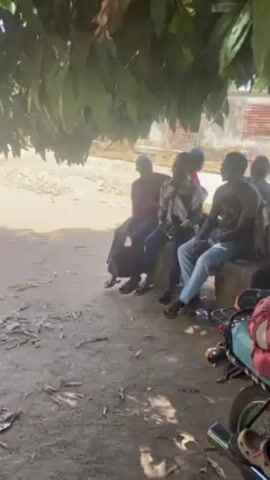 A group of six dark-skinned young men are sitting on a cement bench beneath the shade of a large tree. All are wearing casual clothing. One points a finger in the air while speaking. In the foreground is a motorcycle with items strapped to the seat. The ground is dusty and uneven. There is a building visible behind the figures.
