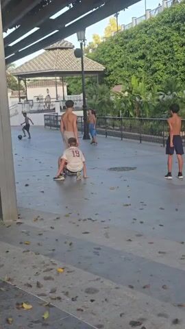 Four boys are playing soccer in a paved outdoor area. The boy on the far left is wearing a white t-shirt and red shorts. The boy in the center is wearing a white t-shirt with the number 19 on it and maroon shorts. He is in a crouching position. To his right is a boy wearing gray shorts and no shirt, and he is dribbling a soccer ball. To the right of him is a boy wearing blue shorts and no shirt. A fifth boy, wearing a white t-shirt and blue shorts, is in the background on the far right. There is a gazebo with a tiled roof and a lamp post in the background, along with a lush green hillside covered in vegetation. Fallen leaves are scattered on the ground.