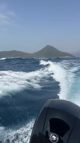 The video shows a first-person perspective of a boat moving through the water. The camera is positioned behind the boat's outboard motor, which is visible in the foreground and is black with a silver logo. The boat is creating a prominent white wake that fans out behind it, indicating movement. The water is a deep blue with moderate waves and white foam. In the background, there is a hazy landscape of green mountains and some buildings scattered along the coastline. The sky is a clear, pale blue. The video appears to be taken on a sunny day, possibly during the day. The audio consists of the sound of the boat's engine and the splashing of water.