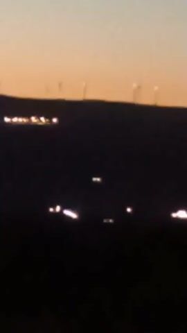 The video is a blurry, dark nighttime or twilight scene. A dark silhouette of a hill or ridge dominates the foreground. On top of the ridge, several distant lights are visible, possibly from buildings or streetlights. Further in the distance, silhouetted against a pale orange and blue sky, are the vertical outlines of several wind turbines. In the middle ground, also in silhouette, are several bright, horizontal streaks of light, suggesting moving vehicles or possibly reflections. Two pairs of distinct, square-shaped lights are also visible in the middle ground, appearing closer than the distant lights on the ridge.