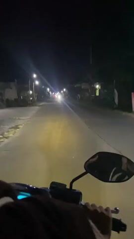This video shows a first-person perspective of someone riding a motorcycle at night. The motorcycle's headlight illuminates a paved road, which is lined with streetlights and occasional houses or buildings. The sky is dark, indicating it is nighttime. The rider's hands are visible on the handlebars, and the side mirror reflects the surroundings. The road appears to have some traffic, as indicated by distant headlights. The overall scene is one of traveling through a semi-urban or rural area at night.