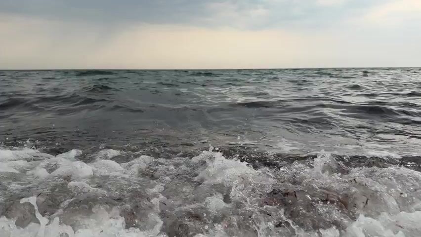 The video shows a low-angle shot of waves breaking on a beach. The water is dark and choppy, with white foam cresting the waves as they reach the shore. The sky above is overcast with a mix of gray and pale yellow clouds. The horizon is visible, separating the sea from the sky. The camera remains stationary throughout the clip, focusing on the continuous motion of the waves. There are no people or man-made objects visible in the frame.