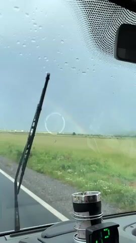 The video shows a rainbow in the distance through a car windshield on a rainy day. The view is of a green field with a few red flowers. A road is visible in the foreground, and there are a few buildings in the far distance. Inside the car, there is an ashtray in a cupholder on the dashboard, and the windshield wipers are visible at the bottom of the frame. The sun glare on the windshield creates a large, translucent circular spot.