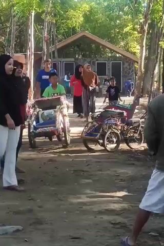 A group of people are gathered outdoors in a wooded area. A boy in a green shirt is standing near a small, colorful cart. A woman in a black hijab and white pants is walking towards the cart. Another woman in a brown hijab and striped pants is standing behind the cart. A man in a blue shirt is walking away from the cart. A boy in a black shirt is standing to the right of the cart. Two trikes are parked to the right of the cart. There is a small building in the background.