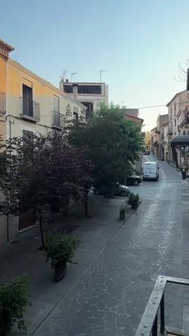 The video pans across a narrow street lined with buildings. The buildings appear to be old, with some featuring stone facades and balconies. Trees are planted along the street, some with dark leaves and others with light green leaves. In the street, a dark-colored car is parked, and a man in a light-colored shirt and dark shorts is walking. The sky is clear and blue, suggesting it is daytime. The lighting indicates that the sun is low in the sky, possibly sunrise or sunset, casting long shadows. There are potted plants visible on a balcony in the foreground. A sign is partially visible on one of the buildings, possibly indicating a business. The overall impression is of a quiet European street scene.