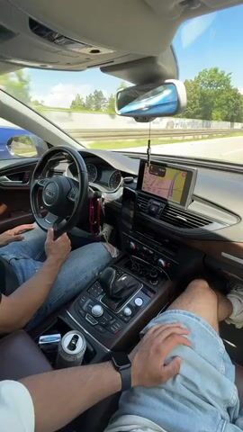 A first-person perspective video captures the interior of an Audi car as it drives on a highway. The driver, wearing jeans, has their hands on the steering wheel and their legs are visible. A passenger, also wearing jeans and a blue short-sleeved shirt, is seated in the passenger seat, with their legs visible and their hand resting on their knee. A can of soda is visible in the cup holder. The car's dashboard displays a GPS navigation system. Outside the car, trees and a blue car are visible, suggesting a sunny day. The video appears to be taken during daytime.