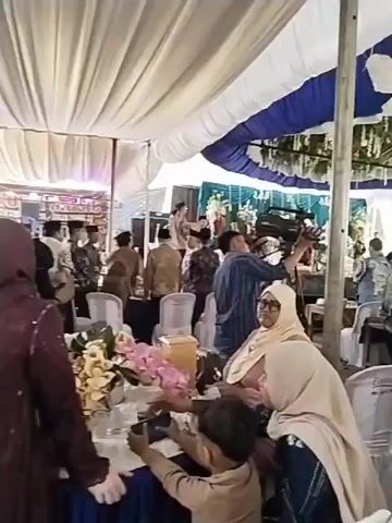 The video shows an indoor event with white and blue tents overhead. Several people are seated at tables, with some standing and milling around. A cameraman is operating a video camera, filming the event. There are floral decorations and what appear to be gift boxes on the tables. The overall atmosphere suggests a celebration, possibly a wedding or similar ceremony.