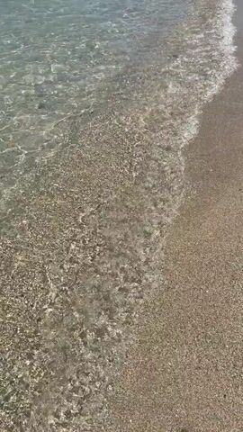 The video begins with a close-up view of shallow, clear water gently lapping onto a sandy beach. The sand is composed of small pebbles and grains. The water is transparent, allowing the sandy bottom to be clearly visible, marked by ripples and the occasional shadow cast by the water's movement. As the video progresses, the camera pans slightly upwards and to the left, revealing a wider view of the beach and the sea. In the distance, mountains form the backdrop under a clear blue sky. Rocky outcrops are visible along the coastline, and a few boats are present in the water. The shoreline is lined with sand, and the waves continue to wash ashore. Further along the beach, a person is standing near the water's edge, facing the sea. The overall scene depicts a tranquil beach environment on a sunny day.