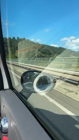 The video shows a view from inside a vehicle, looking out the passenger window. The window reflects the blue sky and green trees. In the side view mirror, a person's hand is visible waving. The vehicle appears to be on a highway, with a bridge and rolling hills in the background. The sky is clear and blue with a few scattered clouds. The lighting suggests it is daytime.