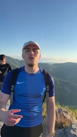 The video shows a young man in a blue Nike t-shirt and a white baseball cap standing on a rocky overlook. He is wearing a black backpack. Behind him, another young man in a black t-shirt and shorts is looking at his phone. The background is a vast mountain landscape with green trees and a clear blue sky. The man in the foreground is speaking to the camera and gesturing with his hands. The lighting suggests it is either sunrise or sunset.