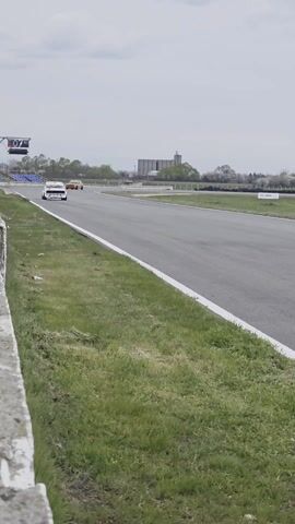 Here's a description of the video:

The video shows a white Lada 2107 race car speeding down a racetrack. The camera is positioned behind a low concrete wall and a wire fence, capturing the car as it approaches and passes by. The car has the number 171 prominently displayed, and it is adorned with racing stickers.

The track itself is asphalt, with green grass and white lines marking the edges. In the background, there are grandstands, buildings, and trees, all under a cloudy sky.

A person's arm is visible in the frame, holding a cell phone to record the car as it races past.