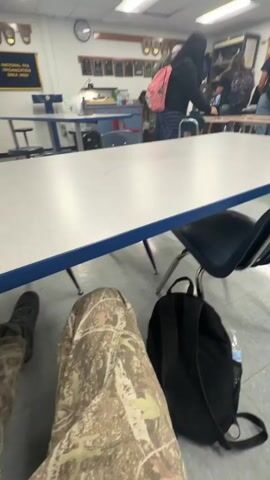 The video shows a classroom environment with several students and tables. In the foreground, a person wearing camouflage pants and a gray sweatshirt is seated, with a black backpack on the floor beside them. Their legs are crossed. A white table with a blue trim runs across the middle of the frame. Behind the table, several students are visible. One student in a black hoodie and striped pants is standing near a table, holding a white object. Other students are scattered around, some with backpacks, in various states of activity. The room appears to be a school classroom or cafeteria, with white walls, fluorescent lighting, and mounted items on the walls. The camera angle is low, looking across the table.