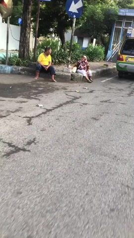 The video shows two people sitting on a curb next to a street. One person is wearing a yellow shirt and blue pants, and the other is wearing a pink and white patterned shirt and dark pants. There is trash on the ground around them. In the background, there is a yellow vehicle and a blue building. A sign with a blue arrow is visible above the people. The scene appears to be outdoors in an urban environment.