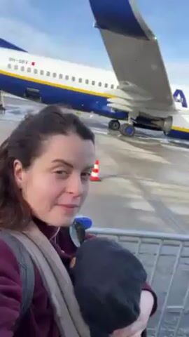 A woman holds a baby in a carrier while standing on the tarmac near a Ryanair airplane. The airplane has a white body with a blue tail and yellow writing. The plane's registration number, 9H-OBY, is visible on the fuselage. The woman wears a burgundy sweatshirt, a grey scarf, and a backpack. The baby wears a dark grey hat. The sky is blue with some clouds.