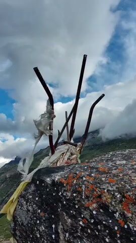 The video pans across a mountainous landscape under a cloudy sky. In the foreground, a large rock is visible with pieces of white and yellow fabric tied to metal rods protruding from it. The camera then pans to reveal a valley with a winding road or river, surrounded by green, grassy slopes and steep, rocky mountains. The clouds cast shadows across the mountains, and patches of blue sky are visible. The overall scene depicts a remote and rugged natural environment.