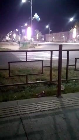 This is a blurry, low-light video recorded at night. It appears to be taken from a sidewalk looking across a street. In the foreground, there is a textured sidewalk and a rusty metal fence with a geometric design. Beyond the fence, there is grass and then a street with a pedestrian crossing. There are streetlights illuminating the scene, and the lights from passing vehicles are visible. In the background, there are buildings and trees. The overall atmosphere is dark and somewhat deserted, with the artificial lighting creating stark contrasts. No people are clearly visible in the frame. The flag on the pole in the middle ground is indistinct due to the low resolution and lighting.