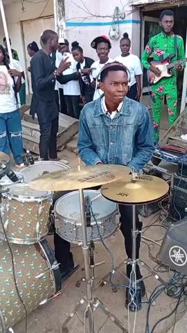 The video shows a drummer playing a drum set in an outdoor setting, with a small crowd of people gathered around. The drummer is a young man wearing a denim jacket, and a pink and white headband. The drum set is decorated with a blue and gold pattern. Behind the drummer, a guitarist in a bright green patterned suit is playing. Several people are standing around, some recording with their phones. The environment appears to be a street in front of a building.