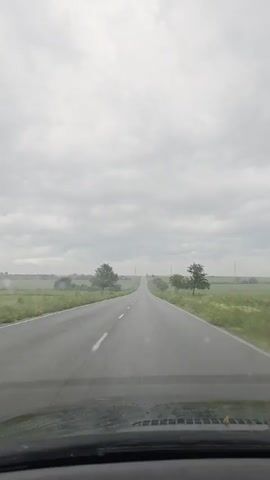 The video captures a first-person perspective from inside a vehicle driving along a long, straight, two-lane asphalt road. The road stretches into the distance, appearing to crest a slight hill. On either side, there are expansive green fields, dotted with occasional trees. Some power lines or utility poles are visible in the far distance, blending into the horizon.

The sky is heavily overcast with a uniform layer of gray clouds, suggesting a cloudy or rainy day. The road surface appears somewhat damp or wet in places, and the lower part of the windshield, visible at the bottom of the frame, shows some water droplets or smudges.

The scene depicts a rural landscape near Iarebichna, Bulgaria. The primary activity is driving, with the camera offering a continuous view forward. There are no other vehicles or people visible, and no discernible text appears in the frame. The overall atmosphere is quiet and serene, characterized by the open road and natural surroundings under a muted sky.