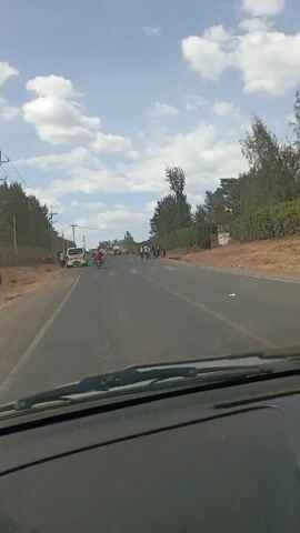 The video shows a car driving down a paved road under a sky with scattered clouds. There are people walking along the side of the road. A sign for the "Ministry of Education, Ndaragwa Comprehensive School" is visible. There are trees and shrubbery along the roadside. Other vehicles, including a van and motorcycles, are also on the road. The camera appears to be mounted on the dashboard of the car, providing a view through the windshield.