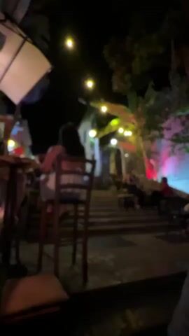 The video depicts a nighttime scene at an outdoor venue, possibly a restaurant or bar, with people seated at tables. The lighting is dim and colorful, with string lights and uplighting illuminating the area. In the foreground, a person is seated at a chair, facing away from the camera. Further back, several groups of people are visible, engaged in conversation or enjoying their surroundings. The atmosphere appears relaxed and convivial. The scene is somewhat blurry, suggesting it was filmed with a mobile device in low light conditions.