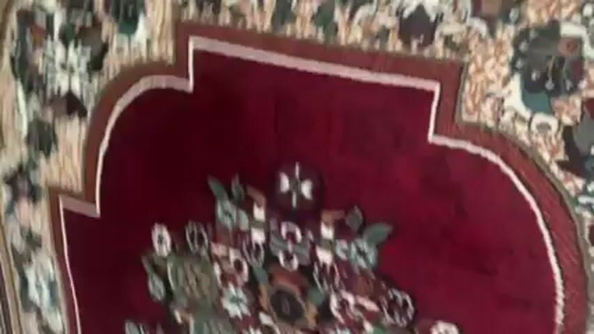 A close-up shot shows a patterned rug with a maroon center section featuring floral designs. The surrounding area has a beige background with floral patterns. The camera moves slightly, revealing more of the rug's design and the overall composition.
