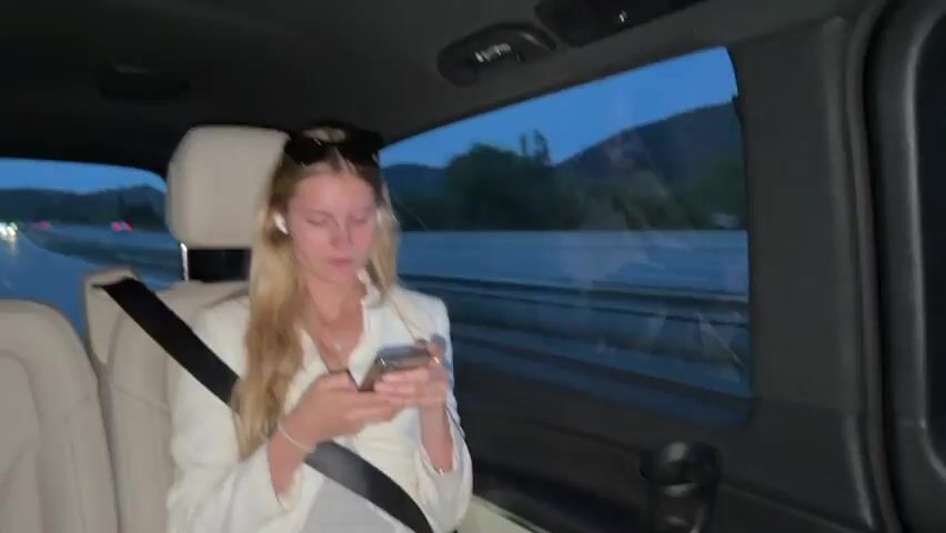 The video shows the view from inside a car, likely a minivan, at dusk. A young woman in a white blazer sits in the backseat, looking at her phone. Outside the window, a highway runs alongside trees and hills. Streetlights are visible. The window is somewhat dirty and has water spots. The camera pans to focus on the window and the passing scenery.