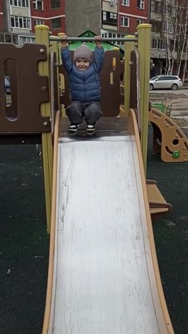 A small child wearing a blue puffer jacket and a gray beanie hangs briefly from a bar at the top of a metal slide in a playground before letting go, squatting on the slide, and then slowly beginning to slide down. Behind the slide is a multi-story red brick building, while the playground surface is green. Another playground structure is visible to the right of the slide, made of tan-colored plastic. The sky appears overcast and gray.
