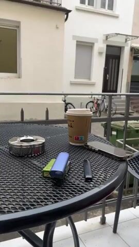 A close-up shot shows a round metal ashtray with cigarettes, two vape pens, a cigarette pack, a mobile phone, and a coffee cup on a black metal mesh table. In the background, two bicycles are parked near trash bins, and the facade of a two-story building with windows and a door is visible. The camera pans upwards to reveal a balcony with an umbrella and potted plants. The video zooms in on the balcony, showing three terracotta pots and a dark-colored pot with a plant.