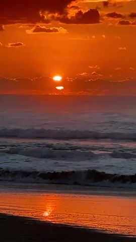 The video shows a sunset over the ocean. The sun is low in the sky, casting an orange glow on the clouds and water. Waves are crashing on the shore, and the sand reflects the sunset colors. The scene is peaceful and serene.