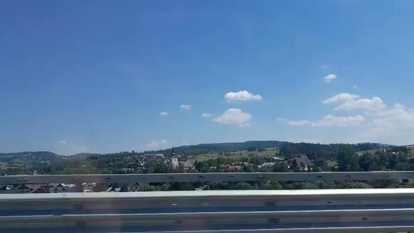 The video captures a daytime view of a rural landscape from what appears to be a moving vehicle, likely on a road or bridge. A metallic guardrail runs across the bottom of the frame, indicating the viewpoint is elevated. Beyond the guardrail, a village or town is nestled in a valley, characterized by a cluster of buildings, some with distinctive architectural features like spires, suggesting a church or similar structure at its center. Rolling hills covered in lush green vegetation and trees form the backdrop, with a clear blue sky dominating the upper portion of the frame, punctuated by a few scattered white clouds. The overall impression is one of a peaceful, scenic countryside on a sunny day.