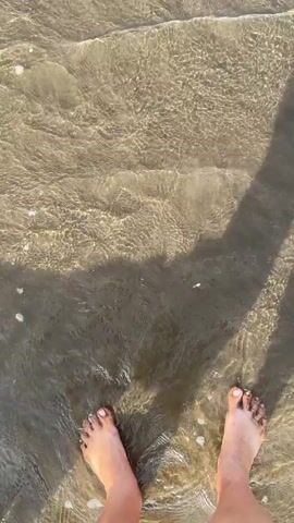 The video opens with a top-down view of a person's feet standing in shallow, clear water at the beach. The sand beneath the water is visible. The person's shadow stretches long across the sand and water from an unseen light source. The camera then pans down and slightly to the right as the person begins to walk into the water. Waves gently lap at their feet and legs. The scene continues with the person walking further into the water, with the waves becoming slightly larger and more foamy. The video ends with a close-up of the person's feet submerged in the water as waves wash over them.