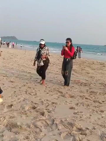 The video shows a group of women walking on a sandy beach with the ocean in the background. The women are dressed in casual attire. Some of them are looking at their phones. The sky is clear and the sun is shining. There are a few other people in the distance on the beach and in the water.