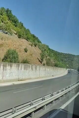 The video shows a view from a vehicle traveling on a highway. The highway is surrounded by a steep, tree-covered hill on the left and a guardrail on the right. The sky is clear and blue. Several cars are visible on the highway, including a black car that passes by the viewer in the foreground. The camera appears to be positioned to capture a view out of the side window of the vehicle.