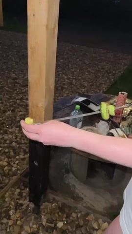 The video shows a triangular cabin at dusk. It's raining. The cabin is decorated with string lights, and it has a deck with stairs leading up to the entrance. There’s a round hot tub next to the deck. The ground is covered with gravel. A hand reaches out and touches a wooden post.