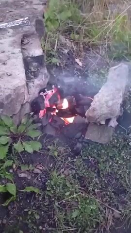 The video shows a small fire burning in a makeshift fire pit surrounded by grass and dirt. The fire pit is constructed from rocks and bricks. Smoke rises from the burning embers, which glow red. Some green leaves are visible around the fire pit. The overall scene suggests an outdoor setting, possibly in a backyard or a rustic area.