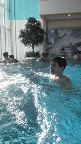 Several people are in a large indoor swimming pool. A young boy is treading water in the foreground. He is looking off to the right. He smiles and waves. Behind the boy are several other people enjoying the water. In the background, there is a decorative tree and a large window that lets in natural light. The pool has a shimmering, turquoise hue. As the camera pans, an adult male with a shaved head is seen neck-deep in the water.